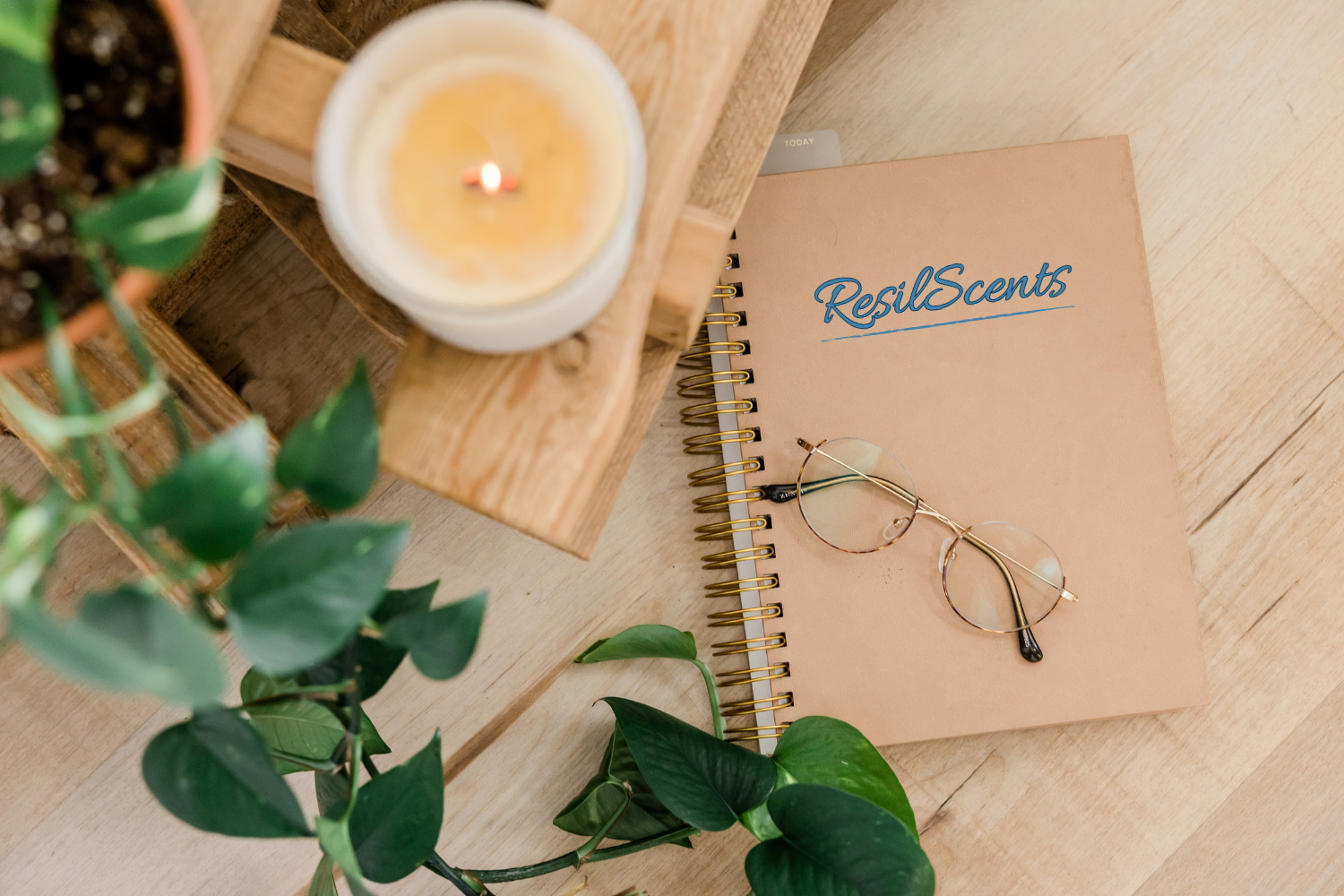“A lit ResilScents™ candle beside a spiral notebook with the ResilScents logo and reading glasses on a wooden desk surrounded by green plants, evoking mindfulness and calm.”