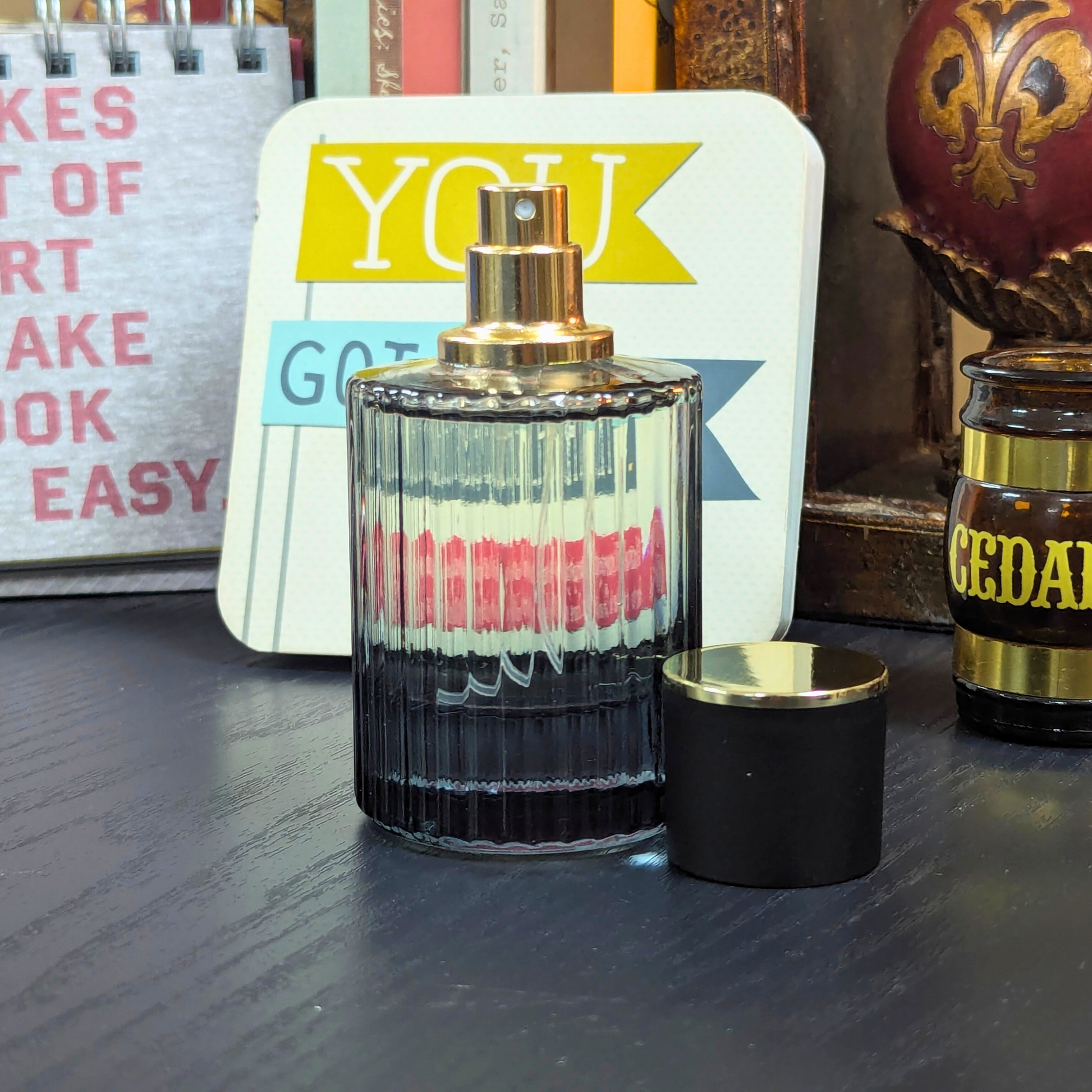 Perfume bottle with gold cap and black base on a wooden surface, with a colorful card and another product in the background.