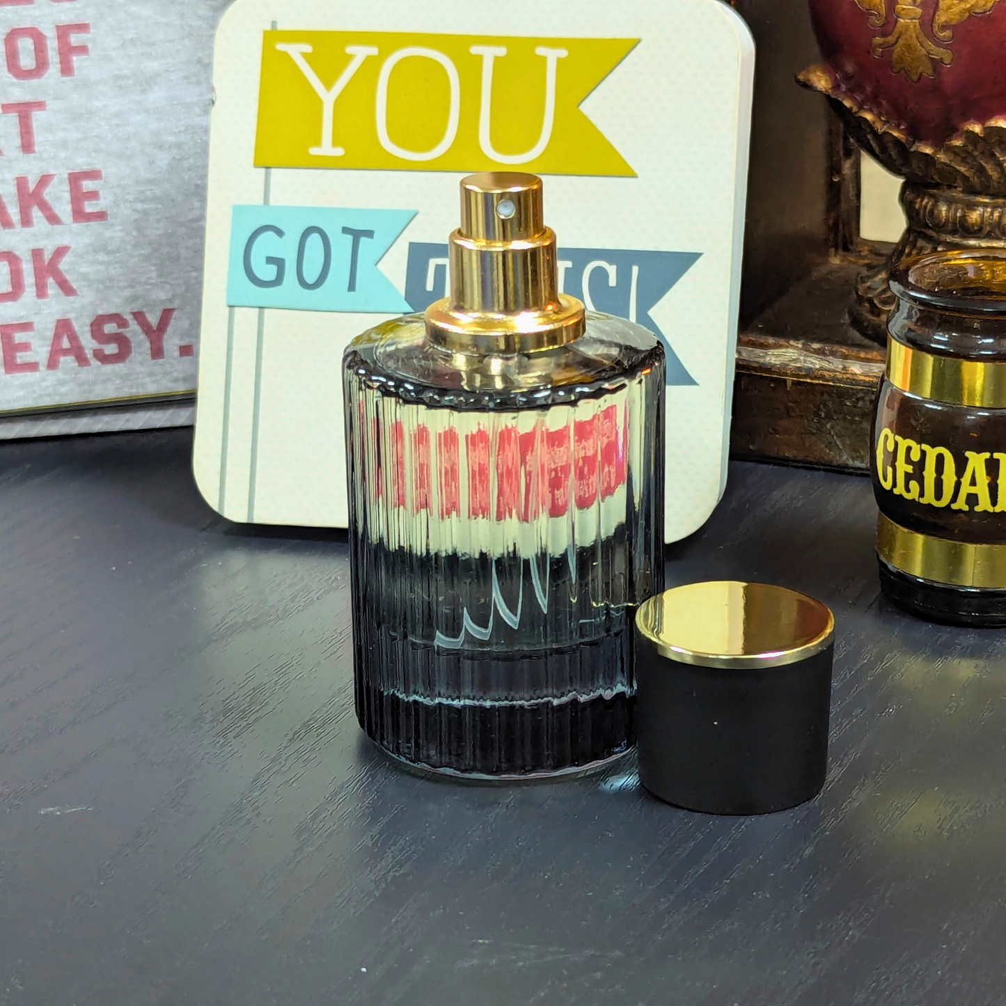 Perfume bottle with gold cap on a dark surface, with a decorative sign in the background.